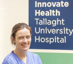 Dr Aoife Doolan smiling and looking straight ahead. Behind her is a large sign that reads: Innovate Health Tallaght University Hospital